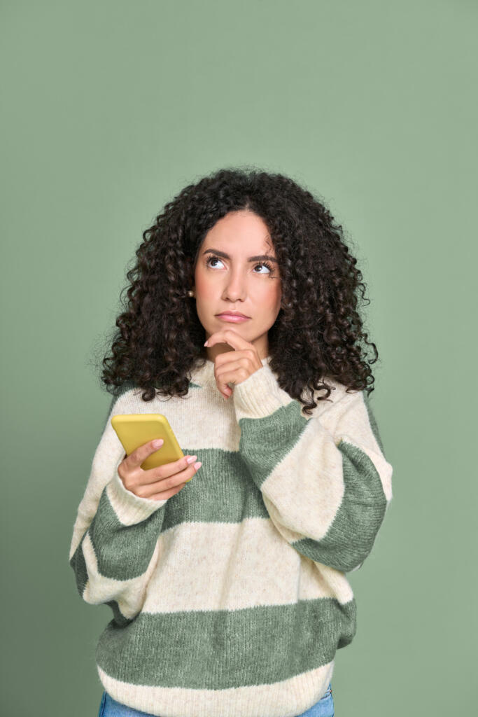 Young thoughtful woman holding yellow mobile phone thinking to herself on a green background.