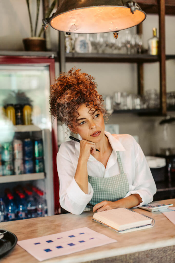 Coffee shop owner looking away thoughtfully while doing her budget in her cafe. Mature business owner calculating her monthly expenses. Businesswoman thinking of new ides for her new business.