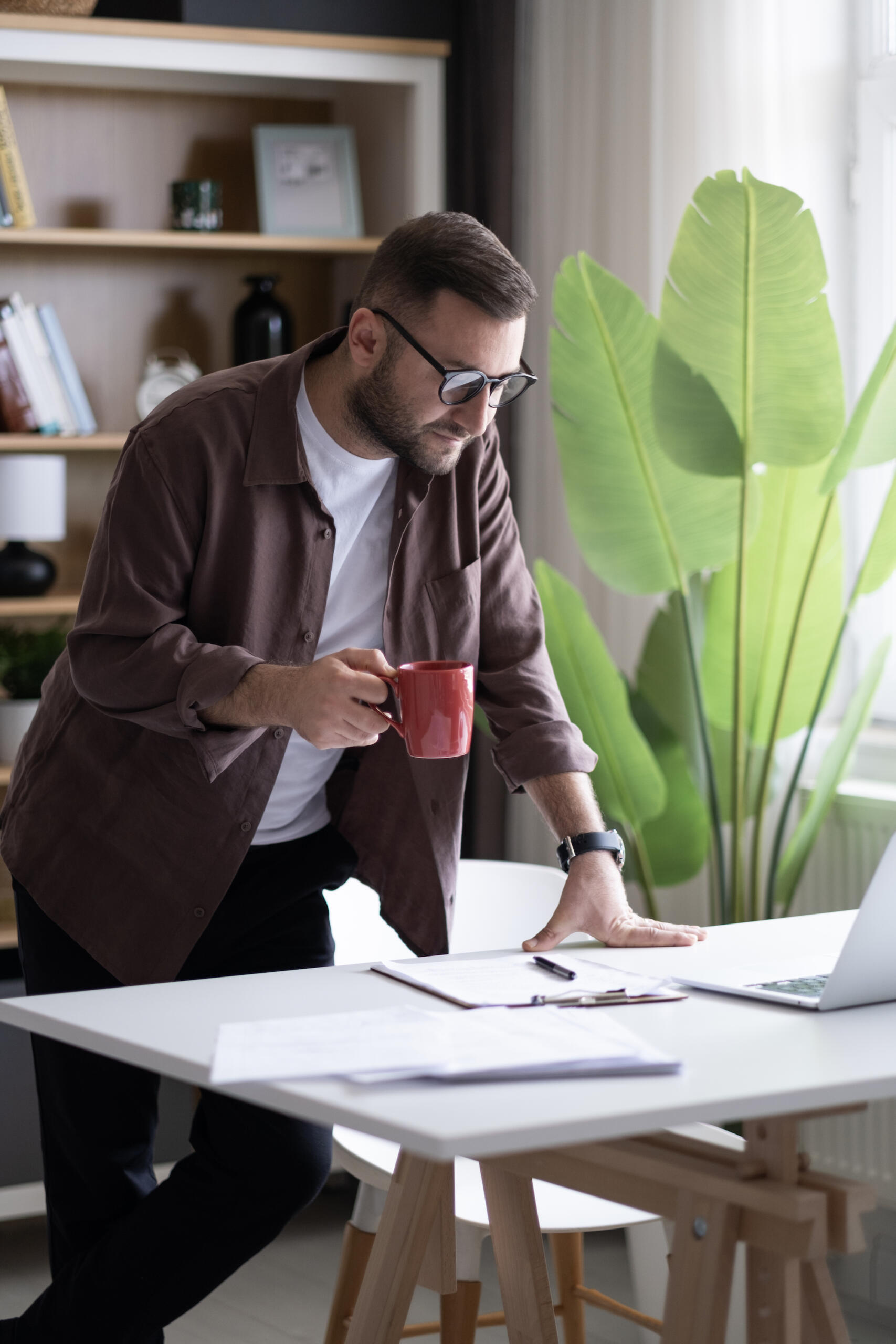 Business owner contemplating his brand name and brand messaging while leaning over a table looking at papers with a cup of coffee in one hand.